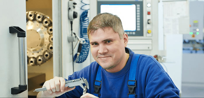 Man in blue overalls holding a silver caliper near metal objects and a computer screen.