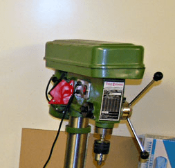 Green metal drilling press with red switch, silver shaft, and black knob against a white wall background.