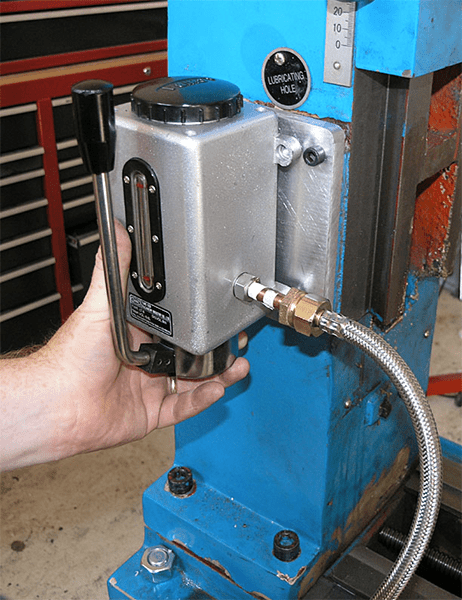 A hand-held lubricating oil canister with a silver body and black knob on top attached to a blue metal machine with a hose.