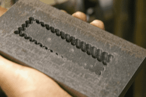 Close-up photograph of rectangular metal object with rough surface and indented text featuring "CNCCOOKBOOK" in block font.