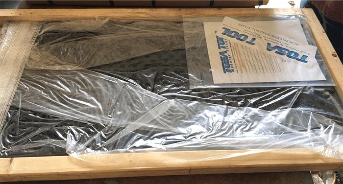 A wooden frame with transparent plastic sheeting, containing a black, rectangular object wrapped in bubble wrap and a white paper with blue "Tool Tusk" text.