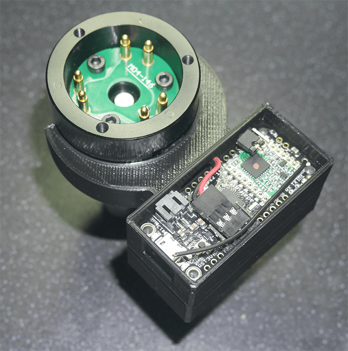 Black cylindrical object with green circuit board and gold pins on dark gray surface.