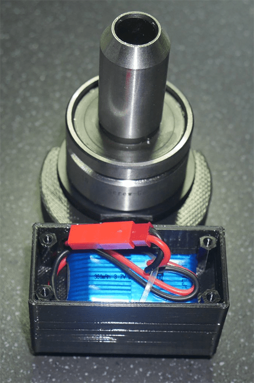 Black cylindrical metal/plastic object with red-blue component attached, situated on gray surface.