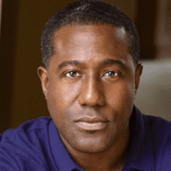 Man with short black hair wearing a purple shirt in a blurred office setting.