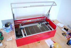 Red laser engraver with glass lid and silver interior on light-colored wooden table.