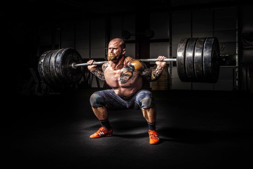 A shirtless weightlifter with tattoos squats with a barbell in a dimly lit gym setting.