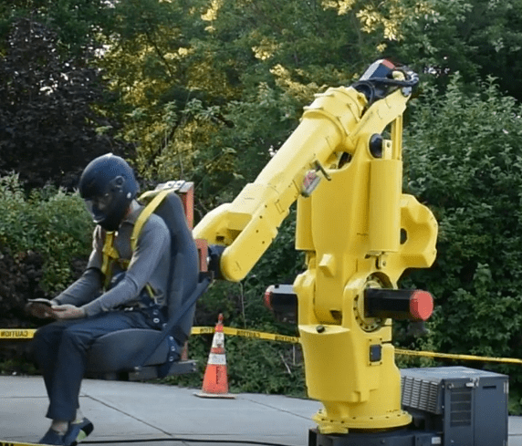 A person sits next to a large yellow robotic arm wearing a black helmet and gray clothing.