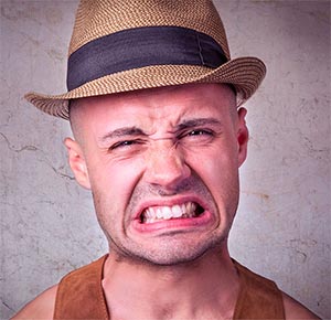 Bald man wearing fedora with black band, displaying an exaggerated grimace face with stubble.
