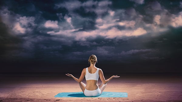 Woman sitting cross-legged on blue mat, meditating outdoors on a cloudy day.