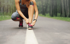 Woman in athletic attire kneeling on a path with tied hands, wearing black tank top and gray shorts, surrounded by trees and grass.