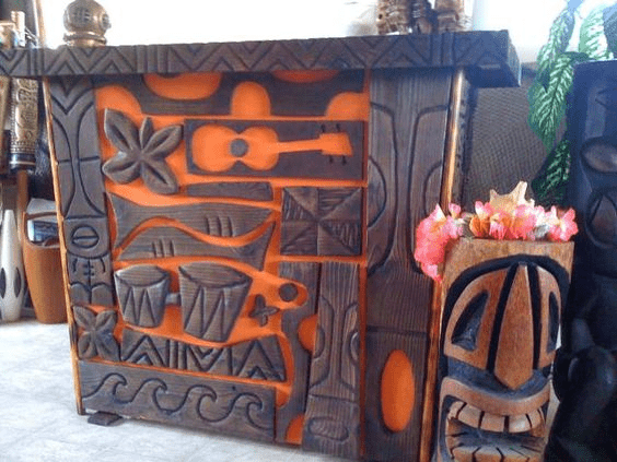 Weathered wooden cabinet with intricate carvings of tiki masks, flowers, and tropical motifs.