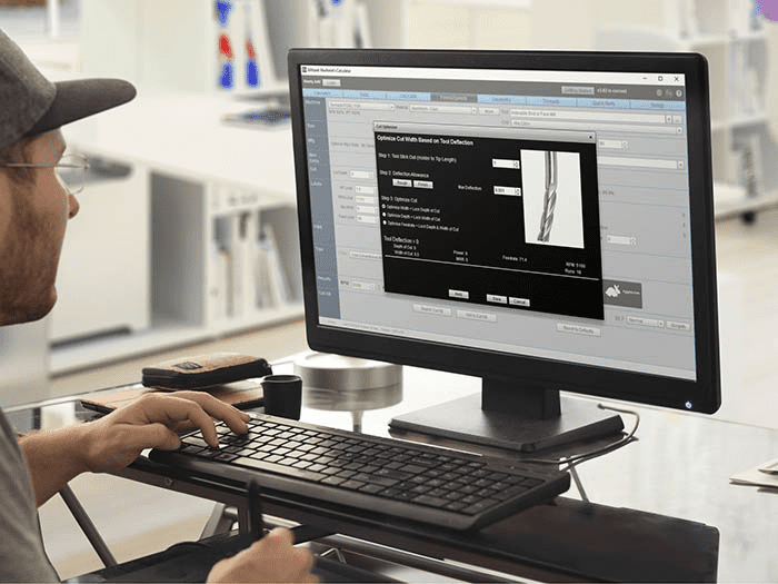 A person sitting at a desk with two computer monitors showing a CAD program displaying a 3D model of metal tubing.