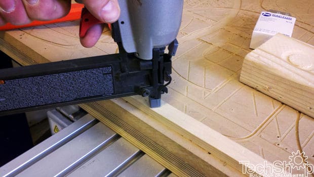 A person holds an air-powered nail gun to a wooden surface on a workbench.