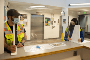 A man wearing a yellow safety vest stands behind a counter holding a piece of paper in a manufacturing or industrial setting.