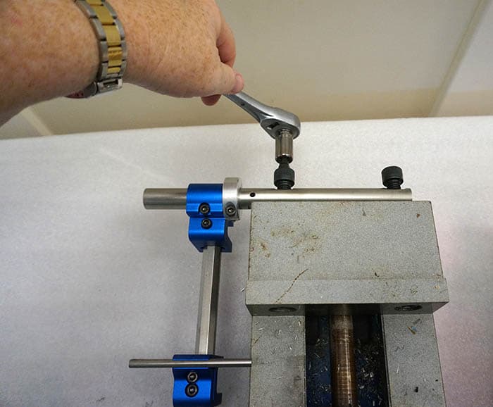 Hand holding a silver wrench in a vise with blue grips, set against an out-of-focus workshop backdrop.