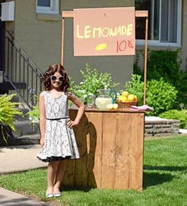 Young girl standing confidently behind wooden lemonade stand wearing white dress with black trim and sunglasses.