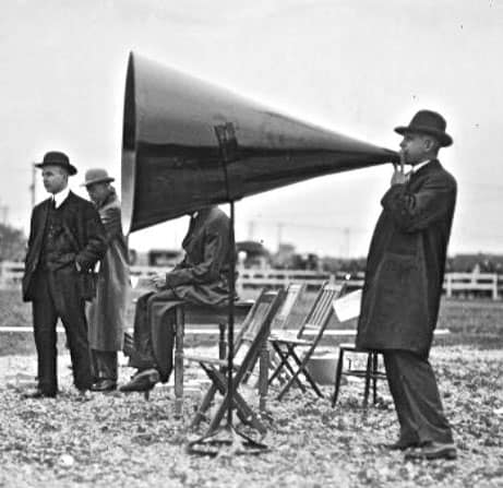 Men in suits, three behind a speaker holding a megaphone, fourth man speaking.