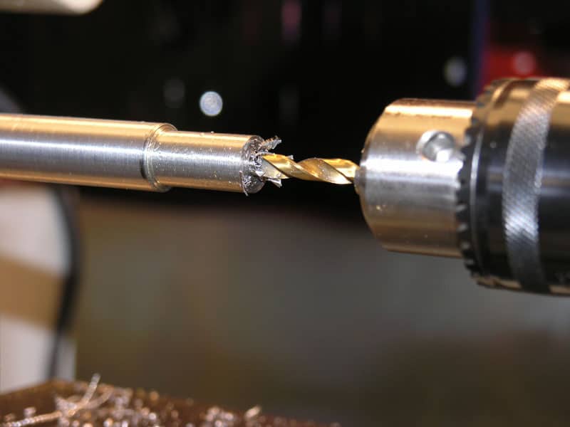 Brass drill bit on drill press, cutting through metal with spiral flute design visible.