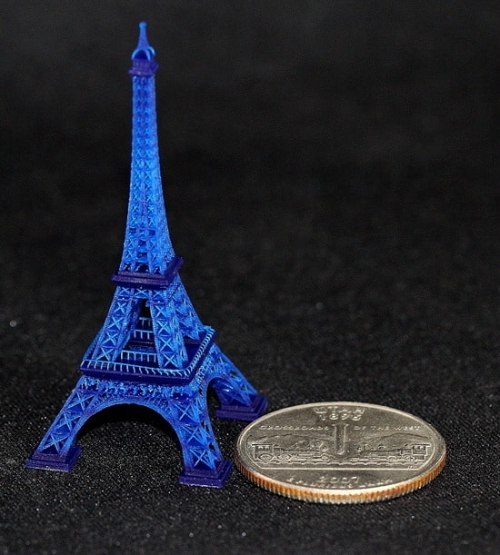 Miniature blue Eiffel Tower next to a quarter coin on a black background.