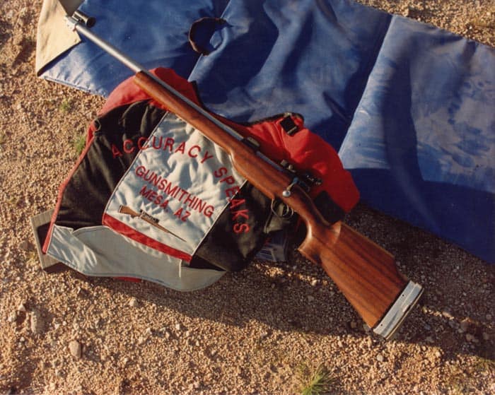 A rifle with wooden stock and metal barrel on a sandy ground next to a bag with red "COURACY SPORTS GUNSMITHING MESQUITE AZ" text.