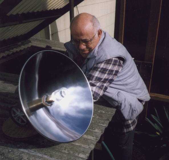 A bald-headed man in glasses examining a large parabolic solar reflector.