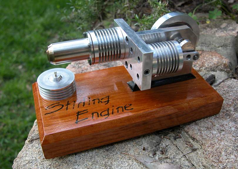 A small metal device with spiral-shaped mechanism and cylindrical component sits atop a light-colored stone surface on a rectangular wooden base inscribed "Stirling Engine".