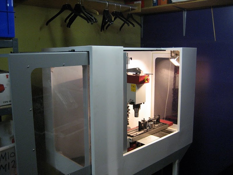 White 3D printer in a cluttered storage room with transparent panels.