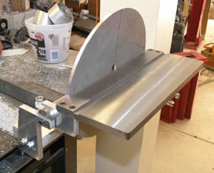 Stainless steel table saw blade guard with curved back plate and rectangular front section, attached by two bolts on the left side.
