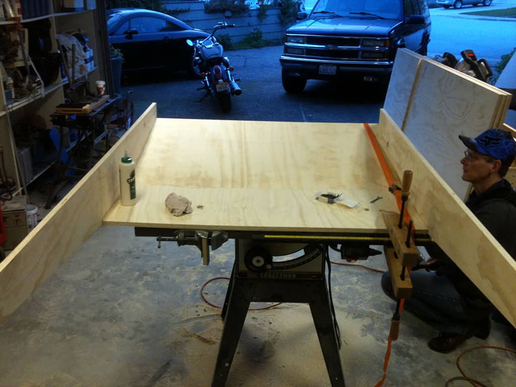 Man kneeling, cutting plywood with table saw in garage workshop.
