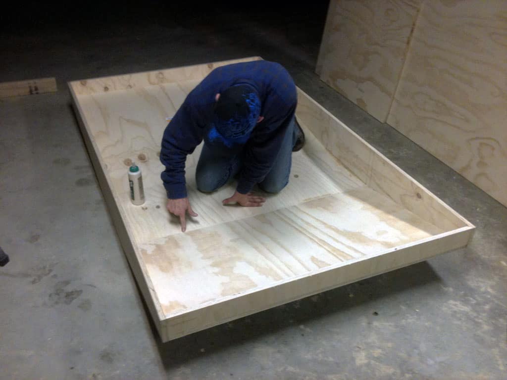 A man kneels on the floor, pointing at a large wooden board with his index finger.