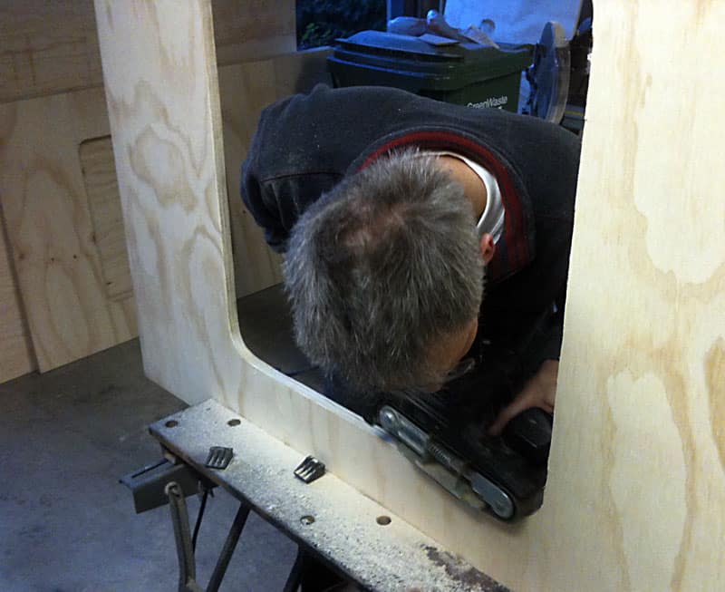 A man sanding the inside of a wooden structure with an electric sander in a workshop setting.