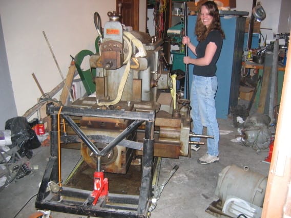 Woman standing beside a large piece of machinery with tools and components in an industrial workshop or garage setting.