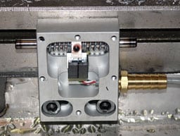 A square metal plate with a cylindrical hole, small red light, and brass-colored rod, attached to a larger metal surface.