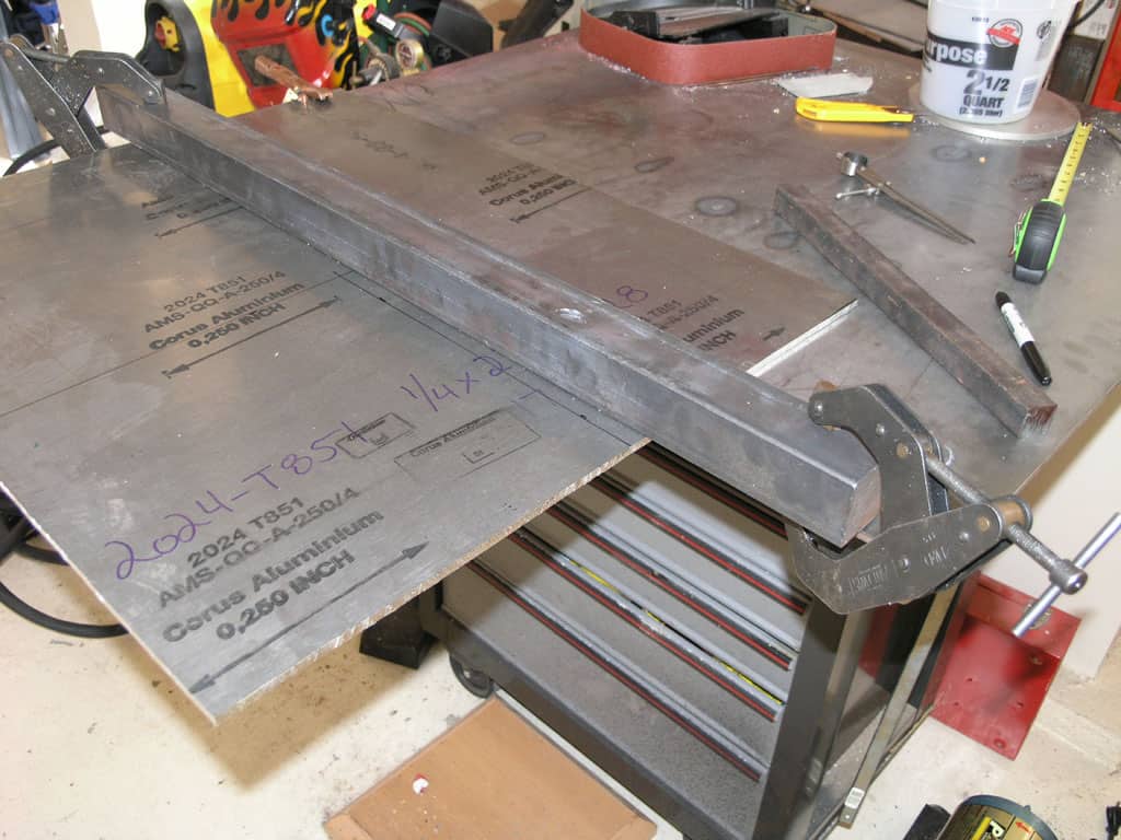 Metal sheet clamped to workbench with metal clamp, featuring a drilled metal sheet with stamped text and numbers in a workshop setting.