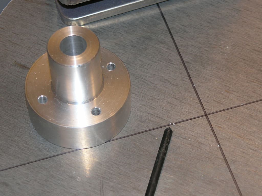 Silver metal object with cylindrical hole and four holes on a gray wooden surface in a workshop.