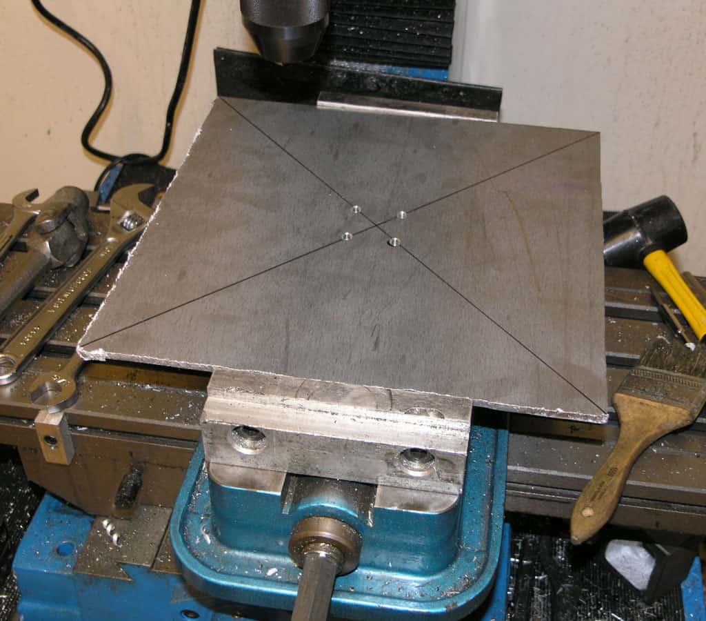 A square metal plate with precision-cut lines on a blue metal base surrounded by tools and equipment in a well-organized workspace.