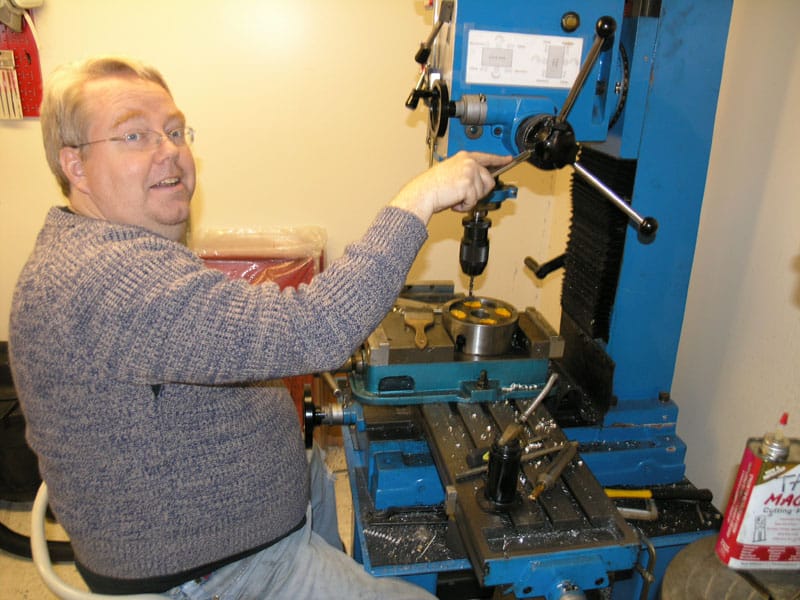 A man in front of a large blue drilling machine, wearing gray sweater and jeans, holding a drill bit.