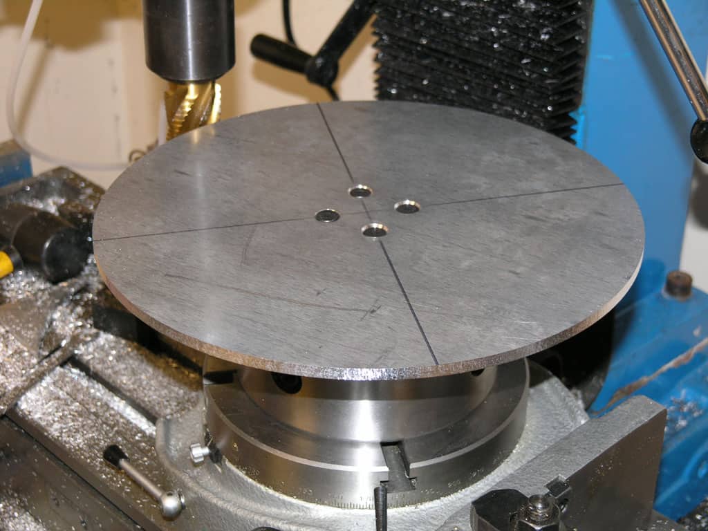 Circular metal plate with four evenly spaced holes mounted on a machine bed.