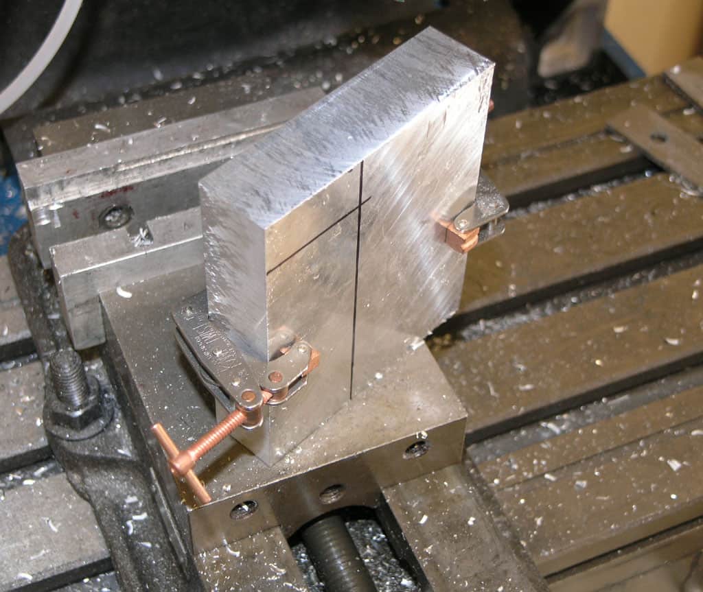 A metal block in a milling machine vise with crosshatch pattern and copper clamps on a cluttered workshop background.