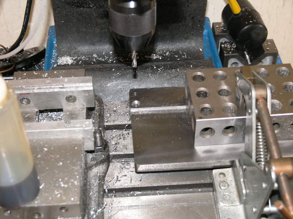 Metalworking machine with drill bit, vise, and workpiece on a blue steel base.