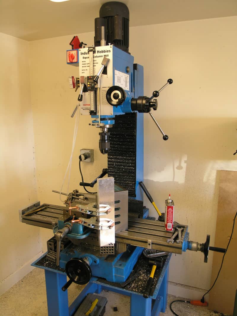 Blue industrial milling machine with tools and parts in a cream-colored walled room.