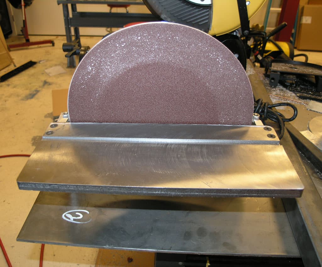 Industrial belt sander on workbench in workshop setting with surrounding tools and equipment.