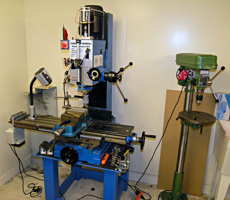 Industrial machines, a blue vertical mill and a green floor-standing drill press, situated in a room with white walls.