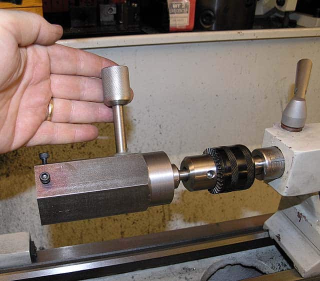 A hand holding a cylindrical object above a metalworking lathe with a large chuck and tailstock on a white background.