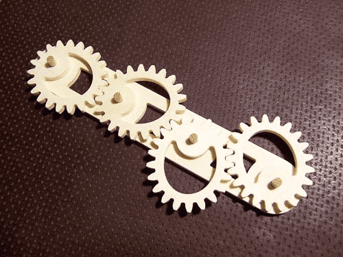 Close-up photograph of three interlocking wooden or plastic gears with a smiley face carved into the center gear.