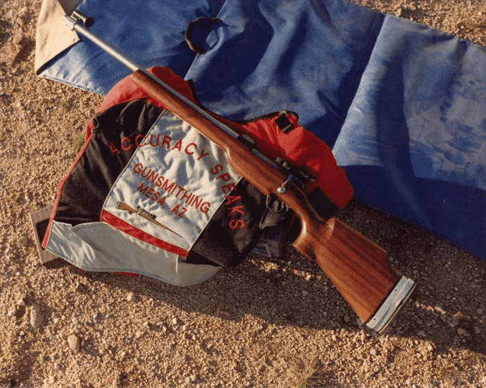 A rifle on the ground with a black and red bag reading "COURACY SPEAKS GUNSMITHING MESA AZ" nearby.