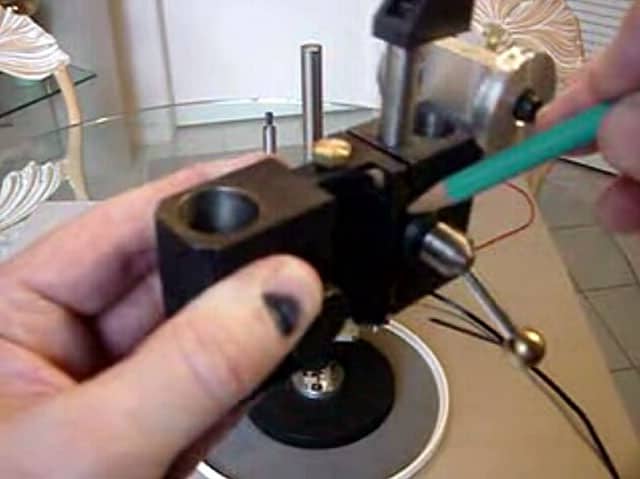 A person's hand holds a black metal object with a hole and green wire on a silver turntable with a blurred background.