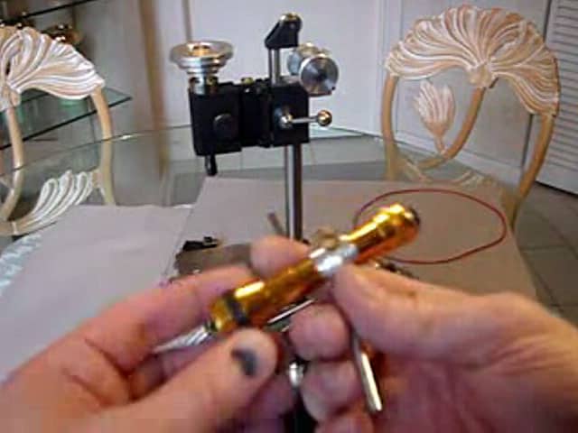 A hand holds a shiny gold tool with a red wire attached to it, set against a metalworking machine background.
