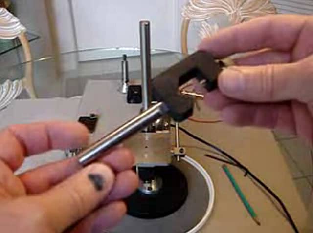 A hand holds a stylus near a turntable on a table, with a metal rod supporting the device.