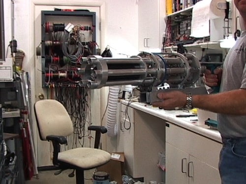 A man stands in front of a cluttered white wall, examining a large cylindrical device with red rings on its surface.